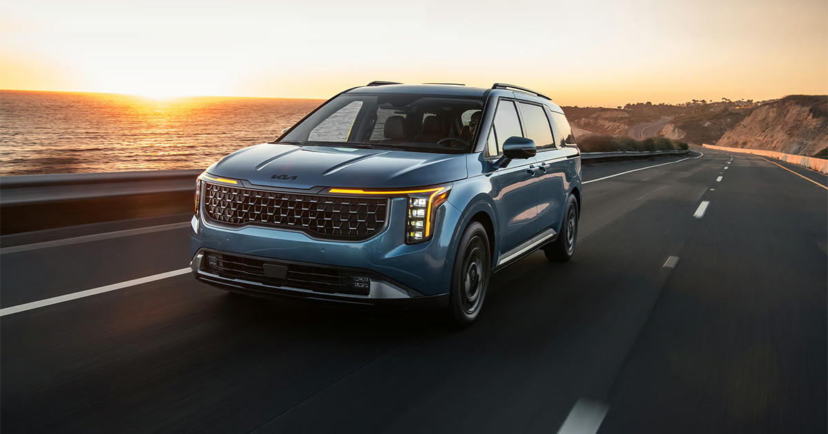 Blue Kia Carnival minivan driving on coastal highway at sunrise