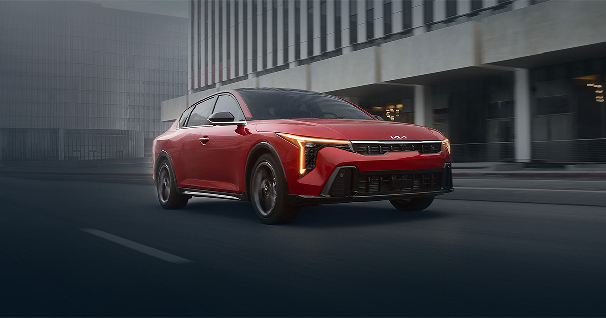 Red Kia K4 sedan driving on city street with modern buildings